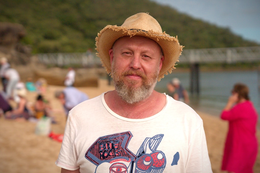 A man in a t-shirt and hat stands on a beach with crowds and a bridge in the background