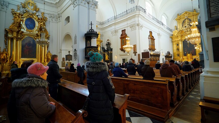 About 50 people in winter coats and hats stand in a catholic church with a gold altar, viewed from the back in a centre aisle