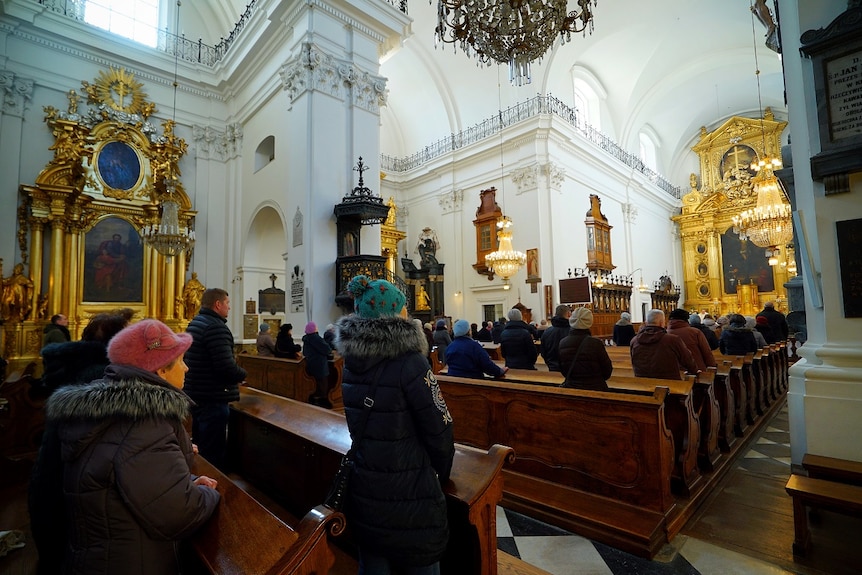 About 50 people in winter coats and hats stand in a catholic church with a gold altar, viewed from the back in a centre aisle