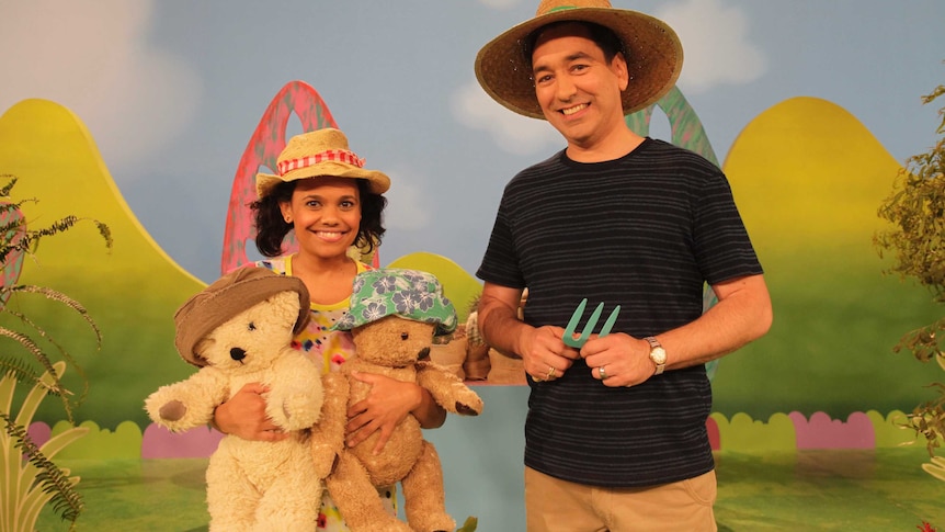 woman and man in hats, woman holding two teddy bears in front of colourful background with green hills