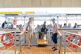 Two people stand on the gangplank leading to a boat that looks like a large tinny holding about 50 passengers.