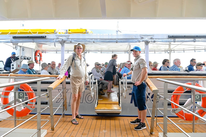 Two people stand on the gangplank leading to a boat that looks like a large tinny holding about 50 passengers.