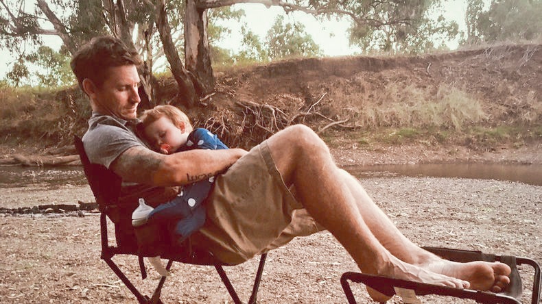 Ryan Murphy cradles his sleeping baby son Wade, as the pair sit on camp chairs beside a river.