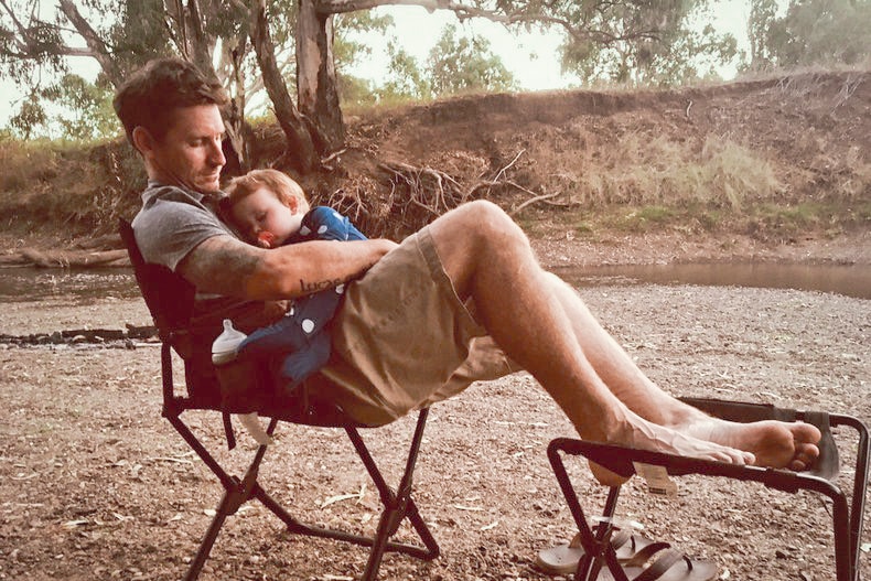 Ryan Murphy cradles his sleeping baby son Wade, as the pair sit on camp chairs beside a river.