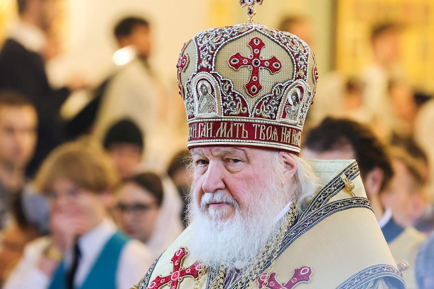 Patriarch Kirill with a white beard looks in the distance as he wears a white hat with a red cross in front of his congregation
