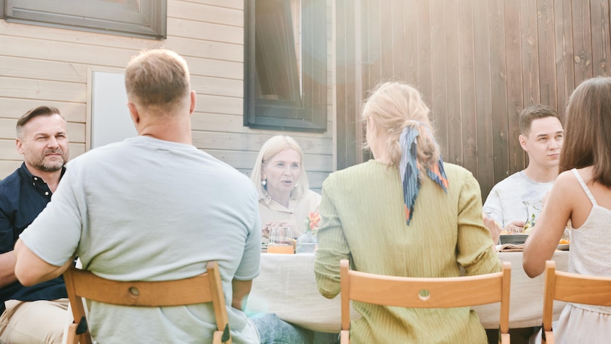 A family of six gather around a table outdoors for lunch, their expressions quite serious.