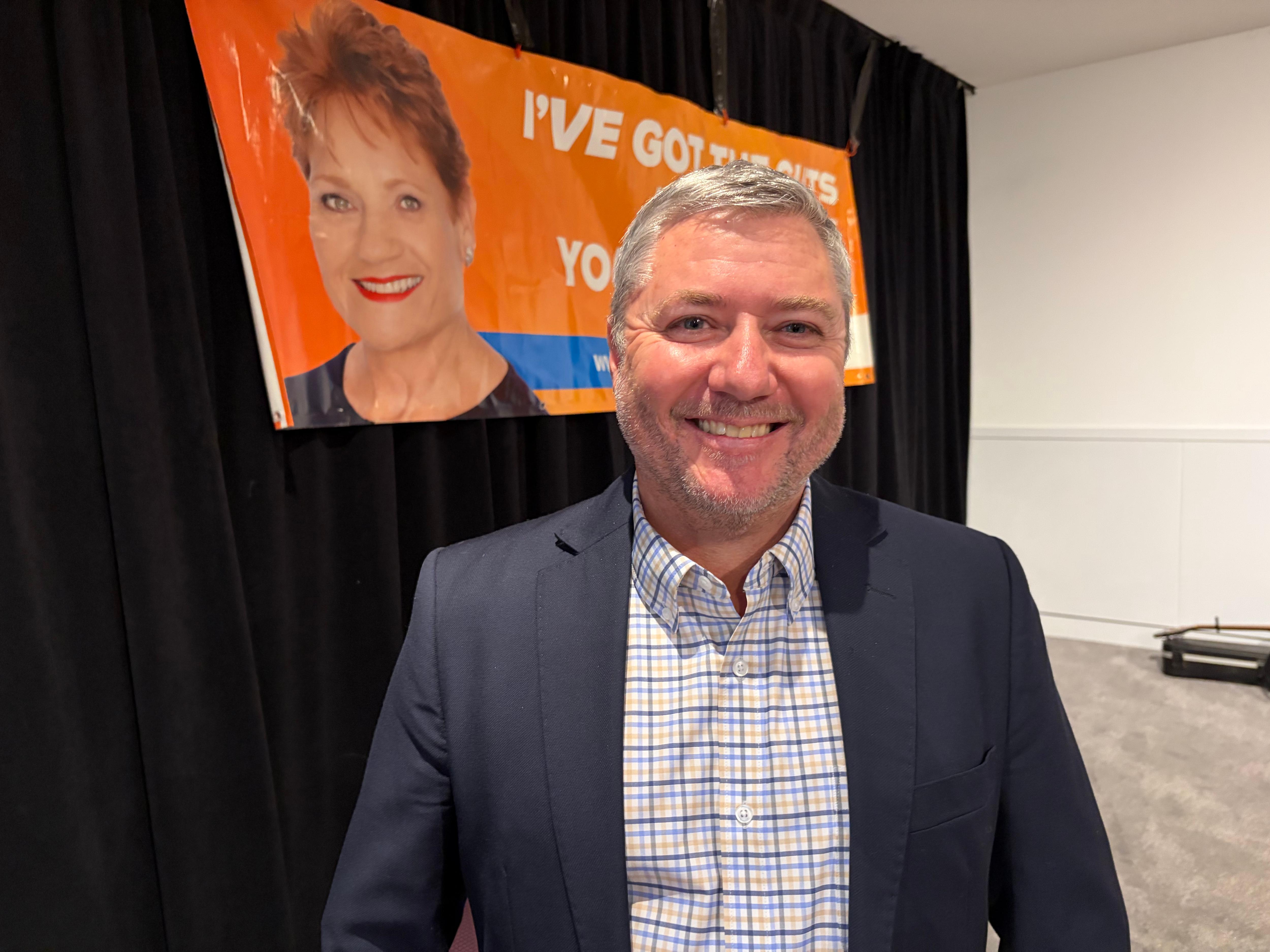 A man smiles at the camera with an orange poster with a photo of One Nation party leader Pauline Hanson on it