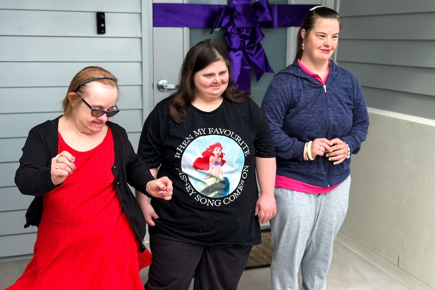 Three women stand outside a new house, all with excited expressions on their faces.