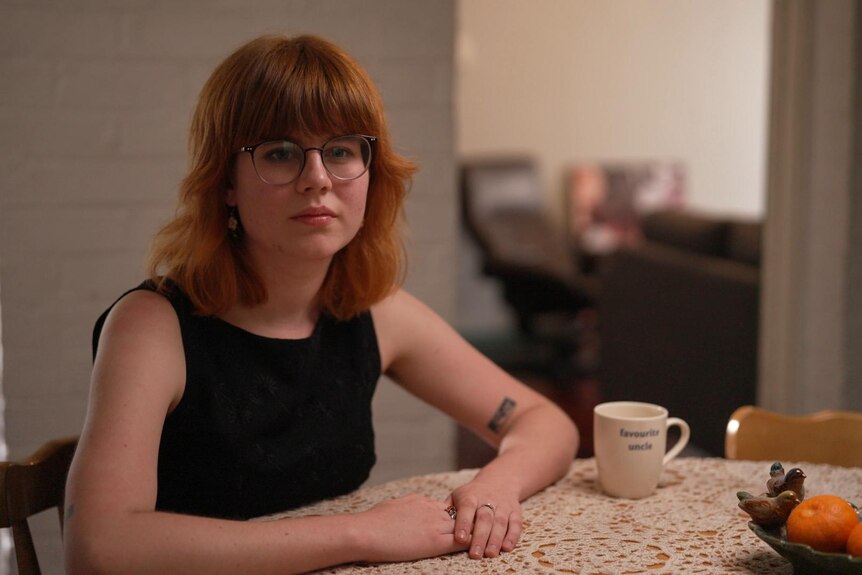 A young woman with red hair and glasses sits at a dining table with a neutral expression