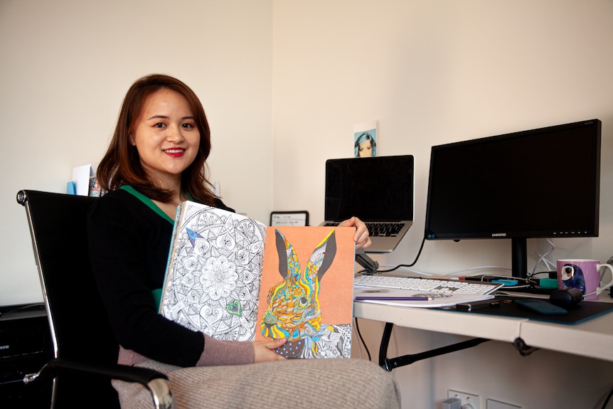 You view a woman of Asian descent sitting down while holding up a colouring book beside two computer monitors.