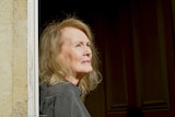 An older white woman with blonde hair leans against a door frame looking into the distance.