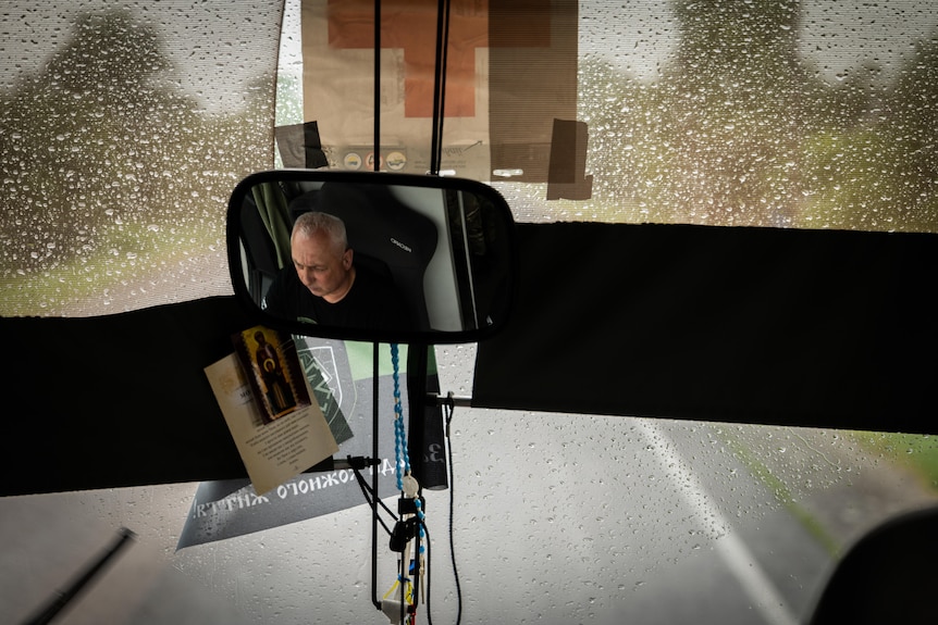A close-up on a rear-view mirror inside a bus shows a driver's reflection. There is rain on the windscreen