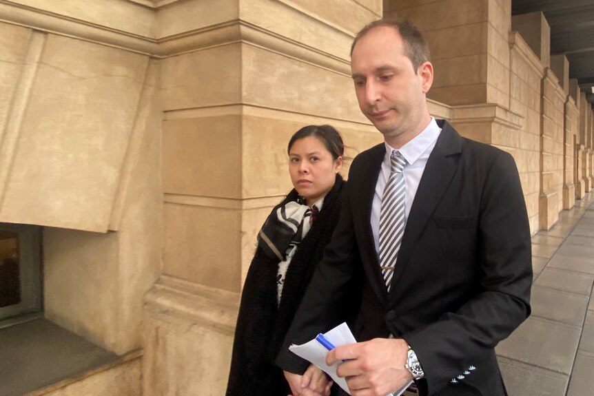A man wearing a black suit looks down as he walks outside a court building. A woman walks beside him