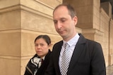 A man wearing a black suit looks down as he walks outside a court building. A woman walks beside him