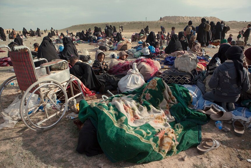 A wide shot shows women and children sitting among blankets and bags of provisions as they prepare to be moved into camps.