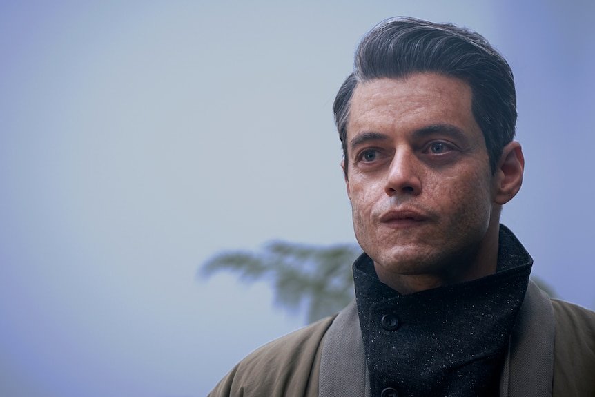 White man with greying hair and scarred face wears a high-collared black and khaki tunic and looks off camera with focus.