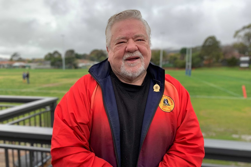 An older man dressed in a rugby tracksuit stands by the field