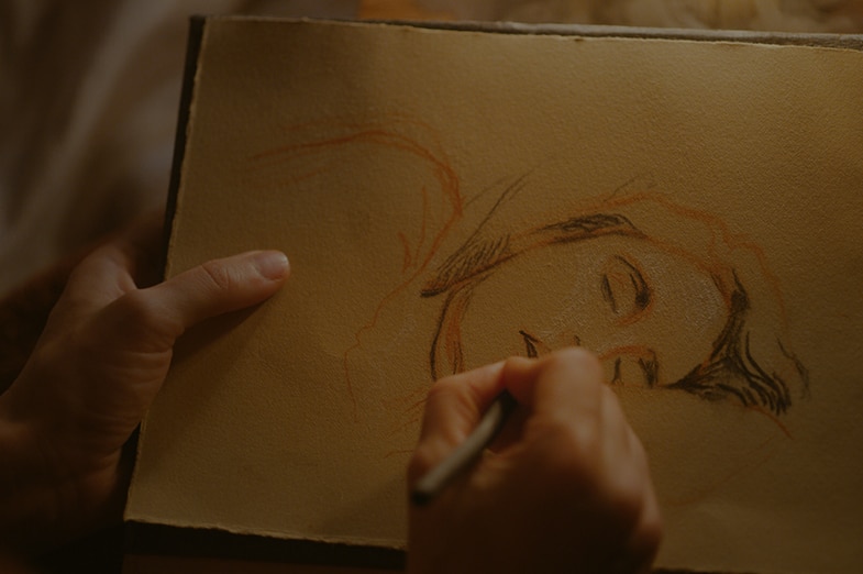 A hand holding pencil draws rough sketch outline of a sleeping woman on texture cream coloured paper.