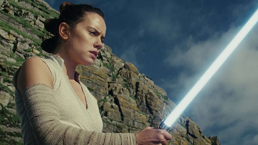 Rey holds a lightsaber, while standing in front of a cliff.