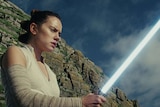 Rey holds a lightsaber, while standing in front of a cliff.
