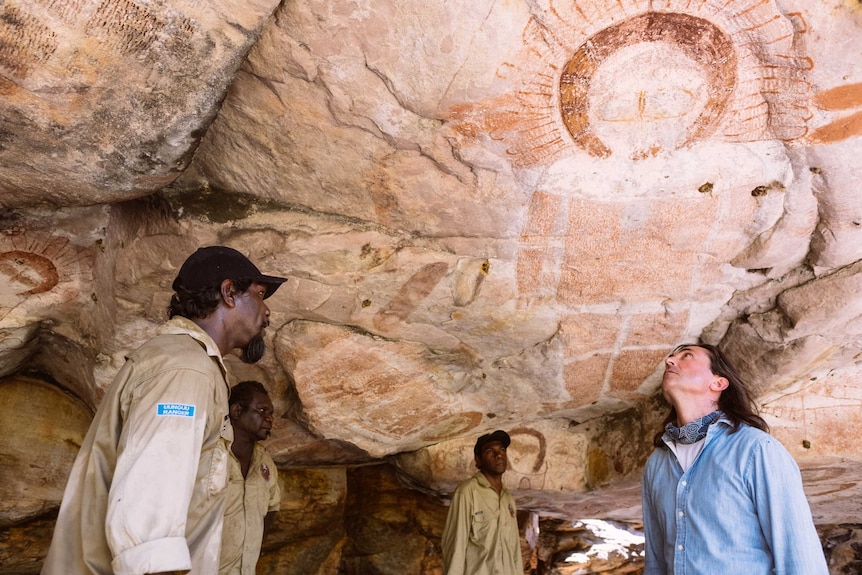 Three Indigenous rangers wearing khaki shirts stand in a cave as a Caucasian tourist in a blue shirt looks at rock art.