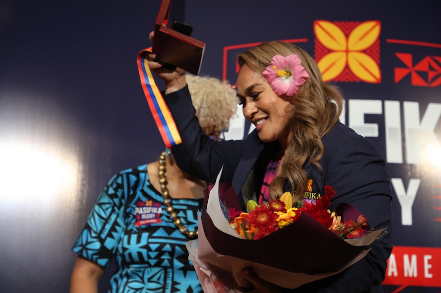 Woman with pink flower in hair wears navy jacket and holds awards as she walks on stage.