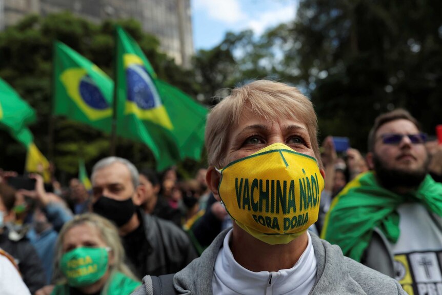 A woman with short blonde hair and yellow mask with a rally crowd behind her waving Brazil flags.