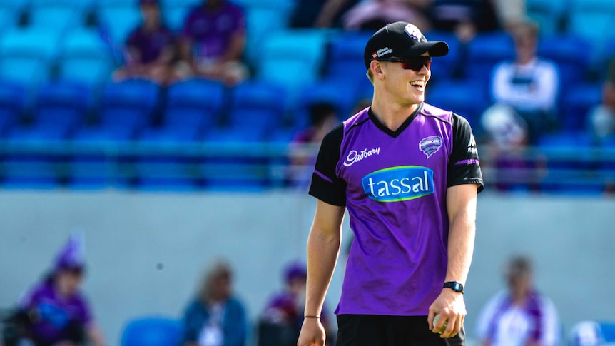 A man in a cap and sunnies smiling, wearing a purple cricket top