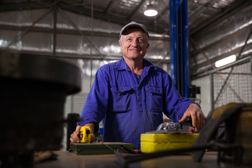 A man, wearing  a cap and a purple shirt, stands in a workshop