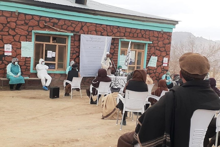 People sit on white chairs in front of a building with information posters stuck on the walls and listen to a speaker.