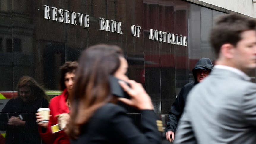 Business people walk past a shiny black sign that reads "Reserve Bank of Australia".