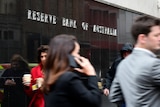 Business people walk past a shiny black sign that reads "Reserve Bank of Australia".