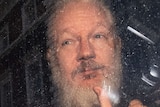 With a silver beard, Julian Assange gives the thumbs up from a police van.