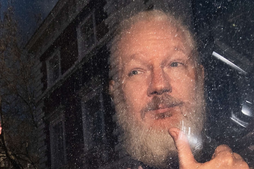 With a silver beard, Julian Assange gives the thumbs up from a police van.