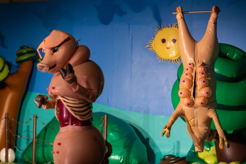 Brightly coloured cartoonish farmyard set. Pig carcass hangs in background. Performer in pig costume stands foreground.