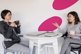 Two women sit across from a table, one talking while the other sips coffee.