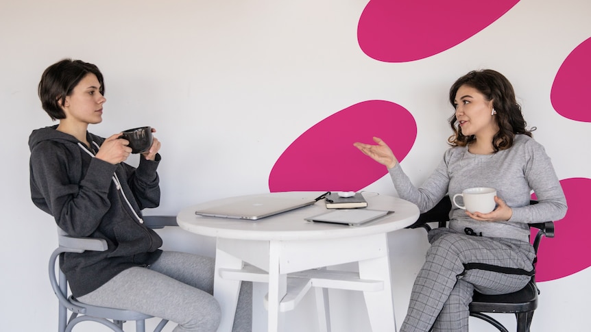 Two women sit across from a table, one talking while the other sips coffee.