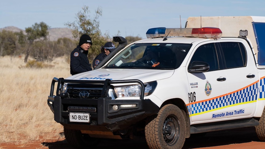 A police truck on a red dirt road, ranges in the background. Two police officers standing outside the car talk to another inside