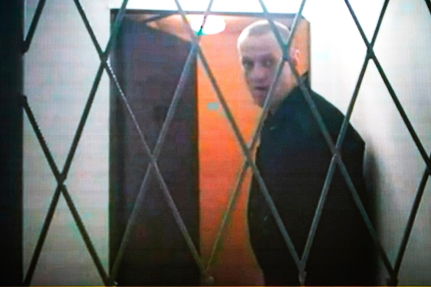 A gaunt-looking middle-aged white man looks through a set of crossed bars.