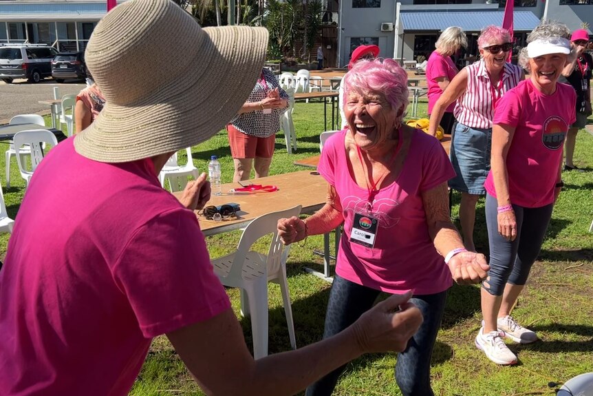 Women dressing in pink T-shirts laughing - a grey and pink haired woman clenching fist leaning forward in full laughter