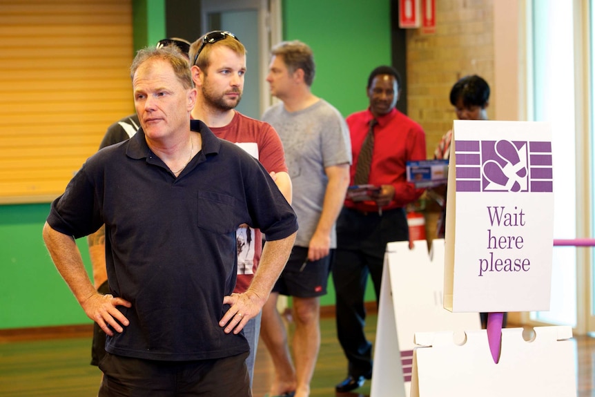 Voters queue up inside the door at a polling station next to a 'Wait here please' sign.