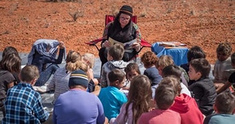 With red sand in the background, a woman sits in a deck chair reading a book to a group of children sitting, facing her.