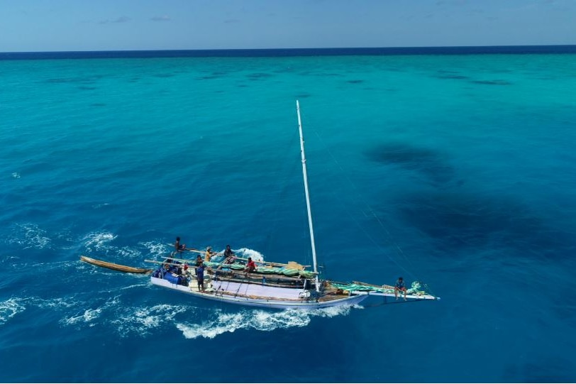 A boat with half a dozen crewmen on board sails through turquoise water