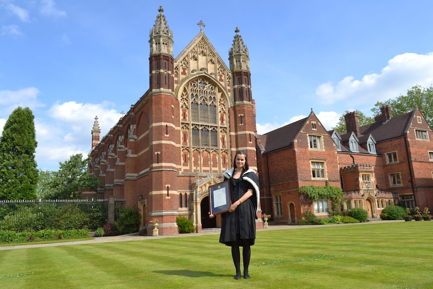 A young woman in a graduation gown, holding a framed certificate in front of a red brick building like a church.