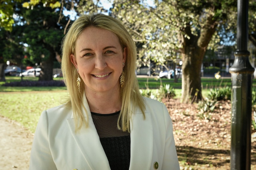 A closeup of a smiling blonde woman in a white jacket and black blouse.