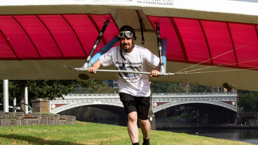 A man runs along grass with a hang-glider type device, river and bridge in background.