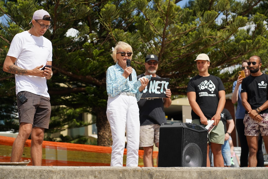 An elderly woman  wearing black sunglasses speaks into a microphone. Man holds sign behind that reads 'no nets'