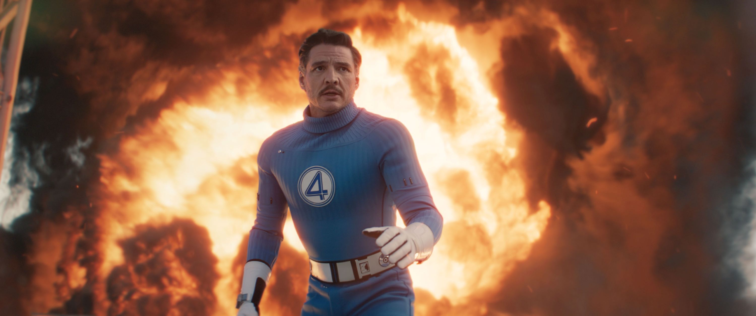 A film still showing a man wearing a blue superhero suit and white gloves and a 4 on his chest in front of a wall of flames