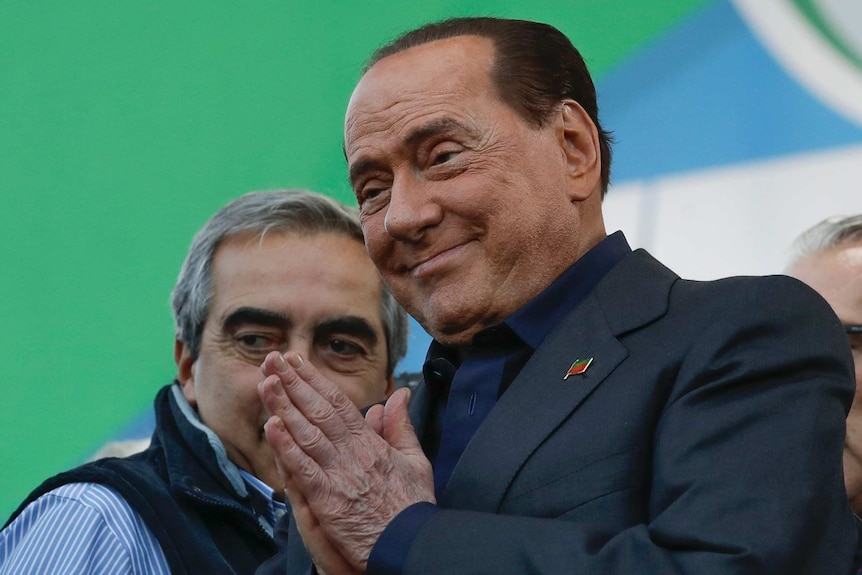 A man in a dark suit presses his palms together and smiles as he poses for a photograph.