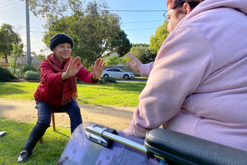 A dance therapist squats with her arms out in front of her as a woman sits in a wheelchair nearby.